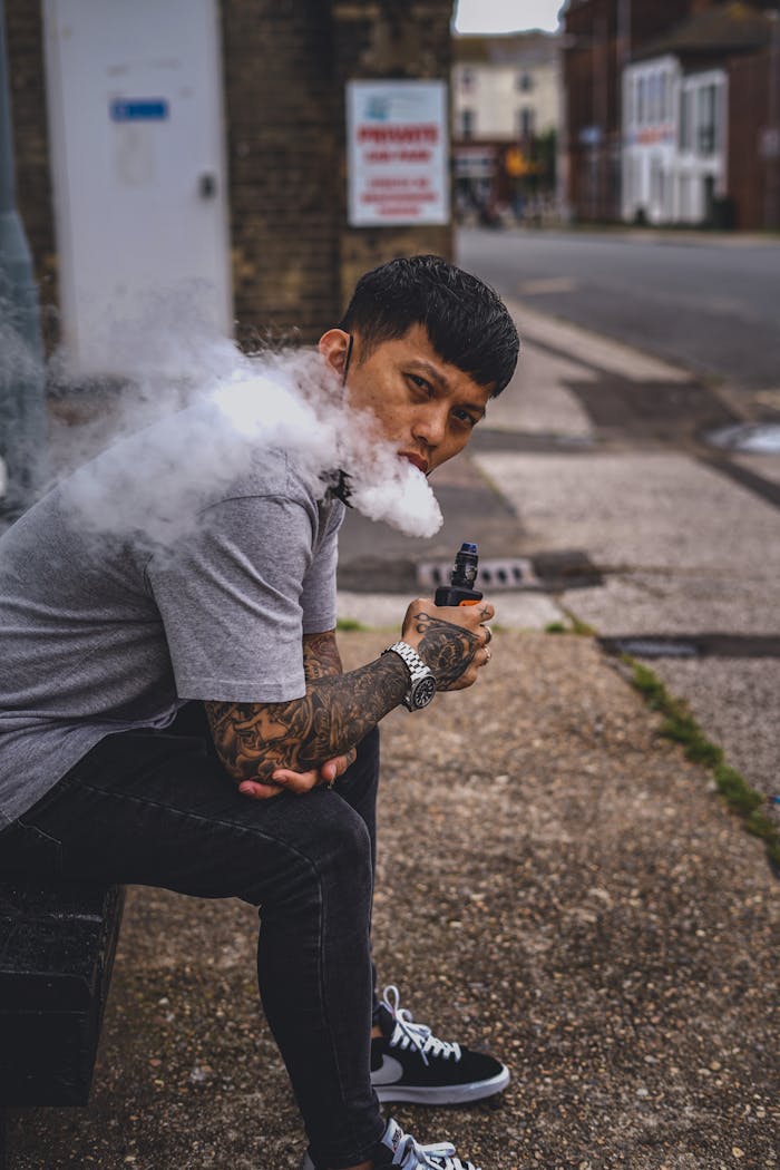 Young man vaping in a city street with tattoos visible, emitting a large cloud of smoke.