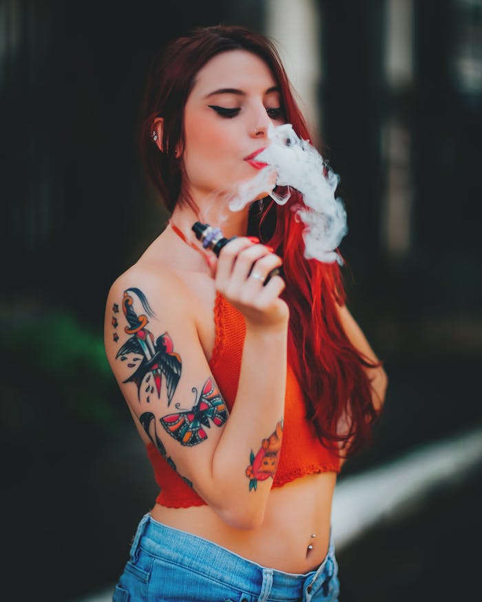services-04 Stylish red-haired woman with tattoos vaping outside on a sunny day.