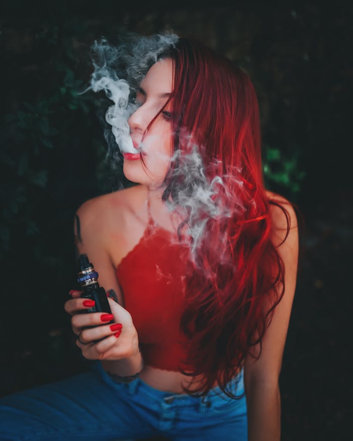 contact-img Young woman with vibrant red hair uses a vape device, surrounded by smoke in an outdoor setting.