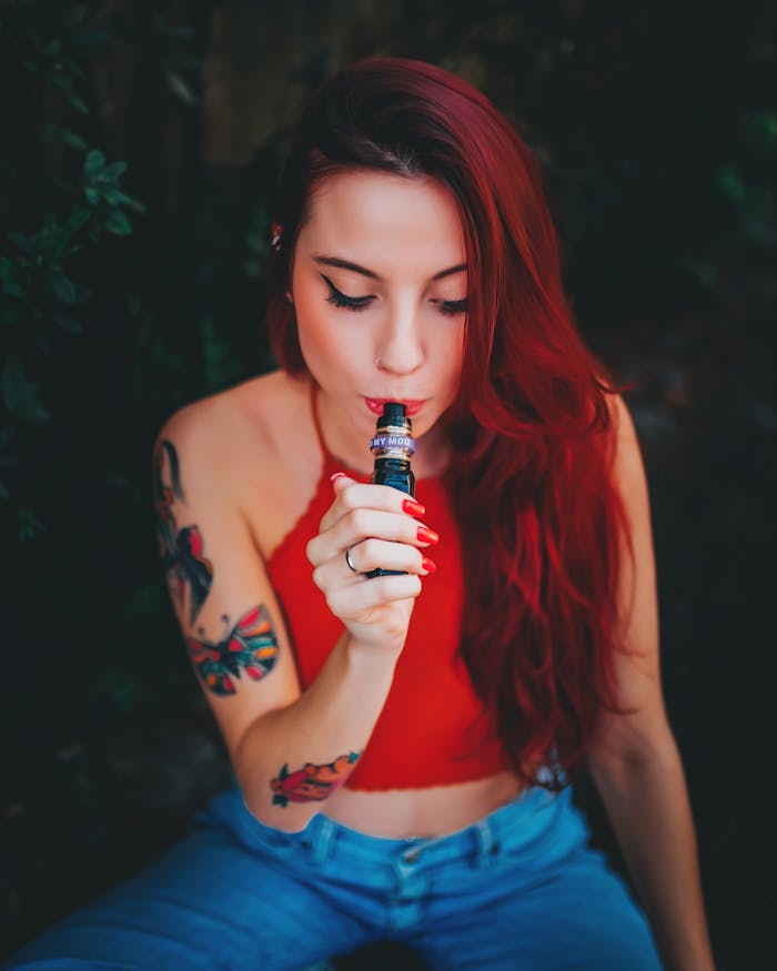 Close-up of a young woman with tattoos and red hair using a vape device outdoors.