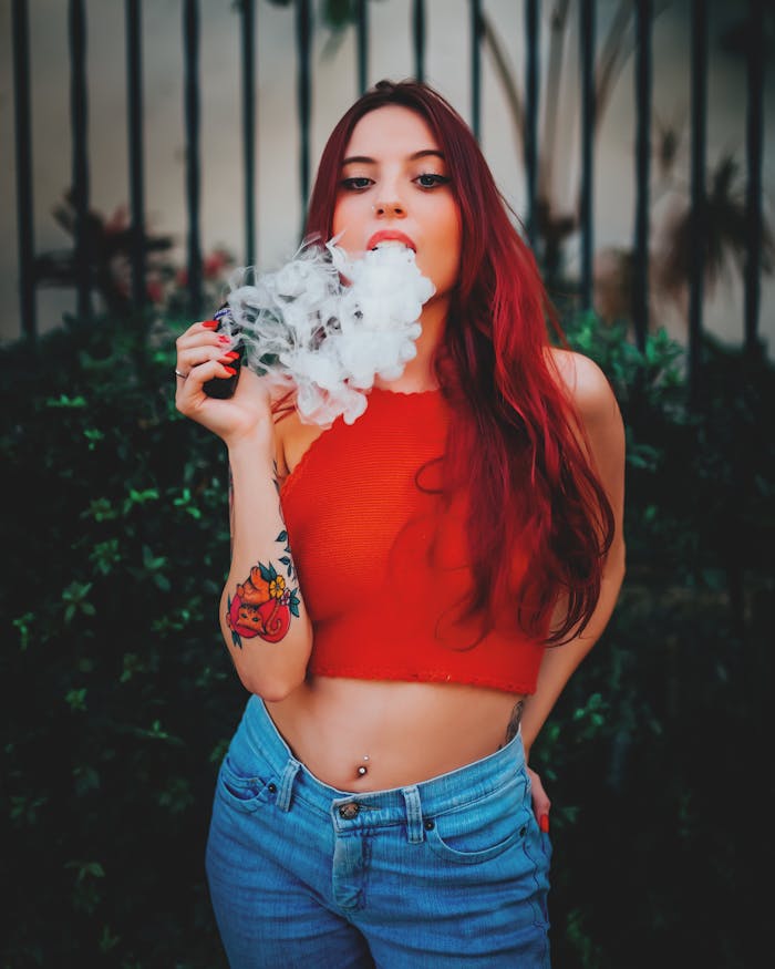 services-01 Young woman with red hair vaping outdoors, wearing a red top and jeans, showcasing tattoos.