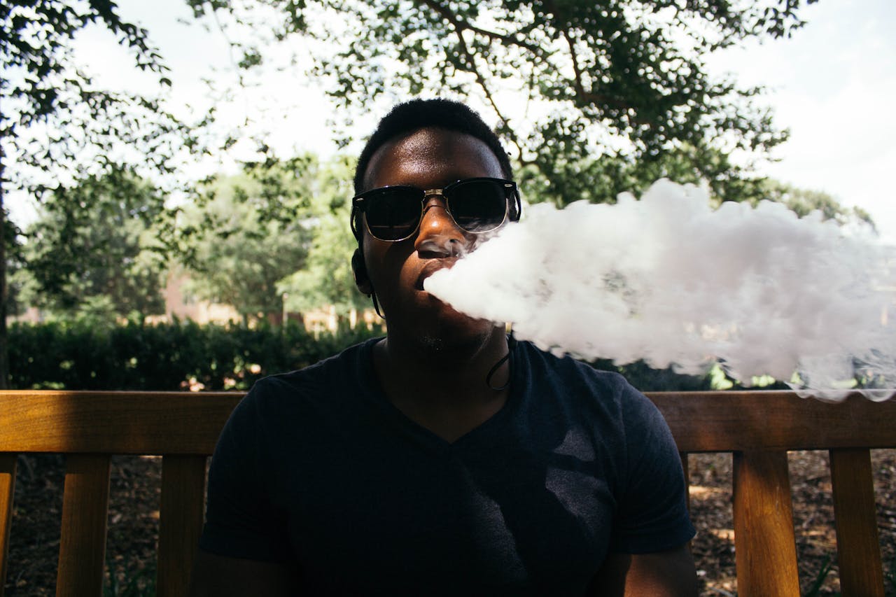 ours-journey A young man wearing sunglasses exhales vapor while sitting on a wooden bench in a sunny outdoor setting.